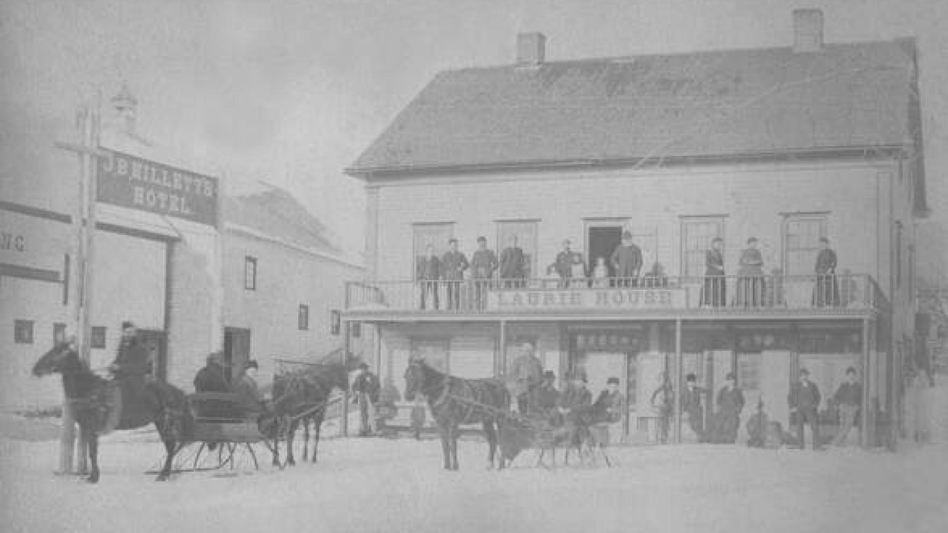 Very early image of the Town Hall building. On the left is a barn with a sign reading JB Millet's Hotel. The Town Hall building has a sign reading Laurie House. There are many people posing on the porches and some horses pulling buggies in the fore ground.