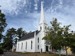 White church with tall steeple against a blue sky, trees beside it.