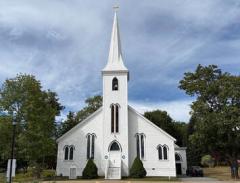 White church with tall steeple, surrounded by trees under a cloudy sky.