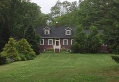 Brick house with dormer windows, surrounded by lush green trees and a large lawn.
